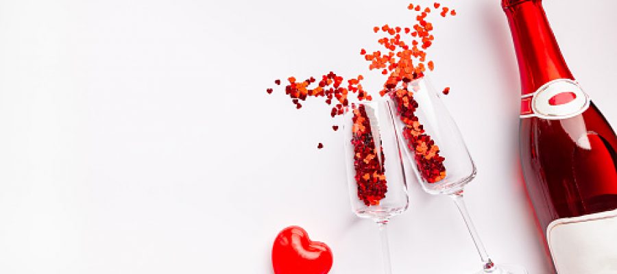 Two champagne glasses with splash of red heart shaped confetti over white background, top view with copy space. Valentine's Day concept